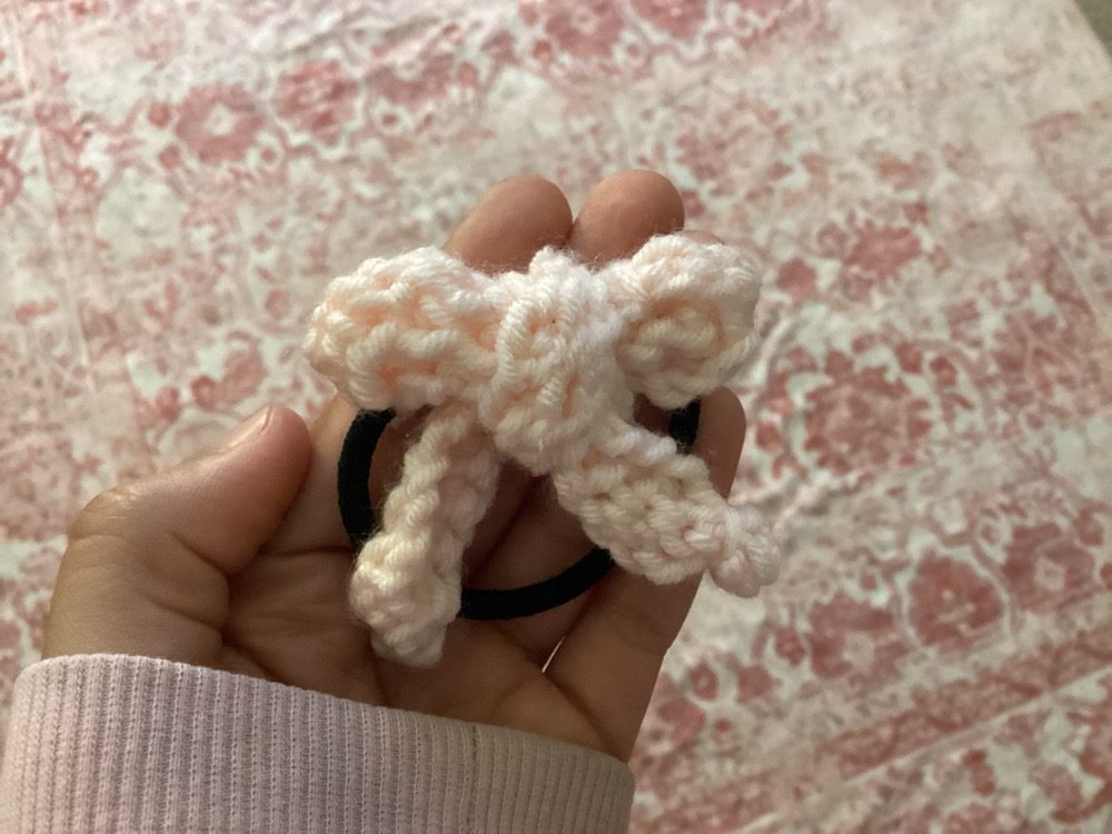 Blush pink crochet bow attached to a black hair elastic, held in a hand