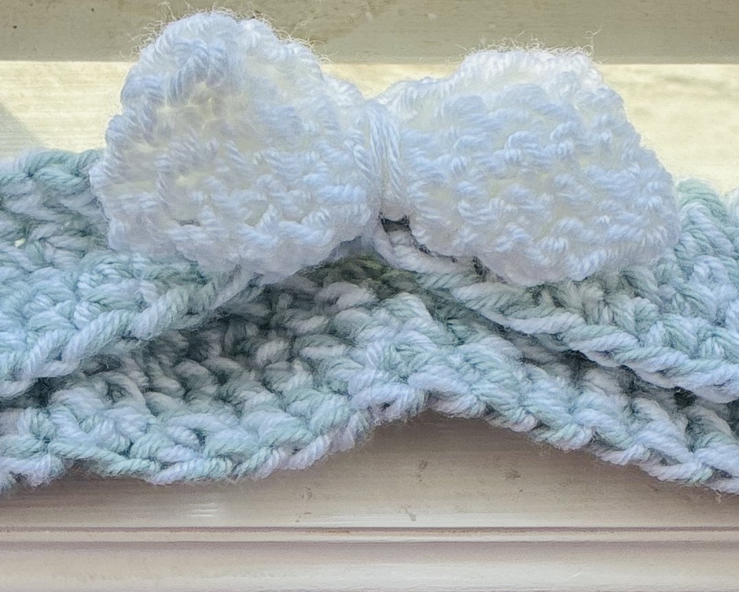 Sage-blue crochet headband with a fluffy white crochet bow, lying on a sunny windowsill