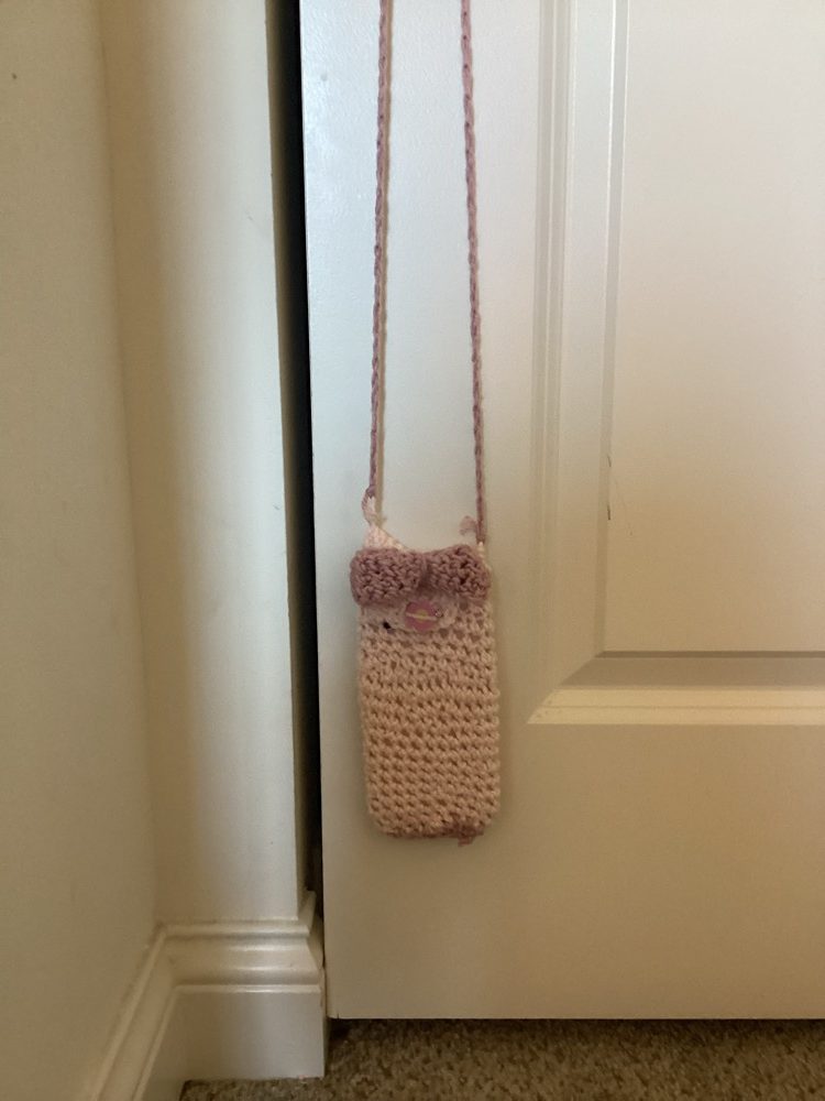 Pale pink crochet phone bag with a mauve bow and button, hanging from a long strap