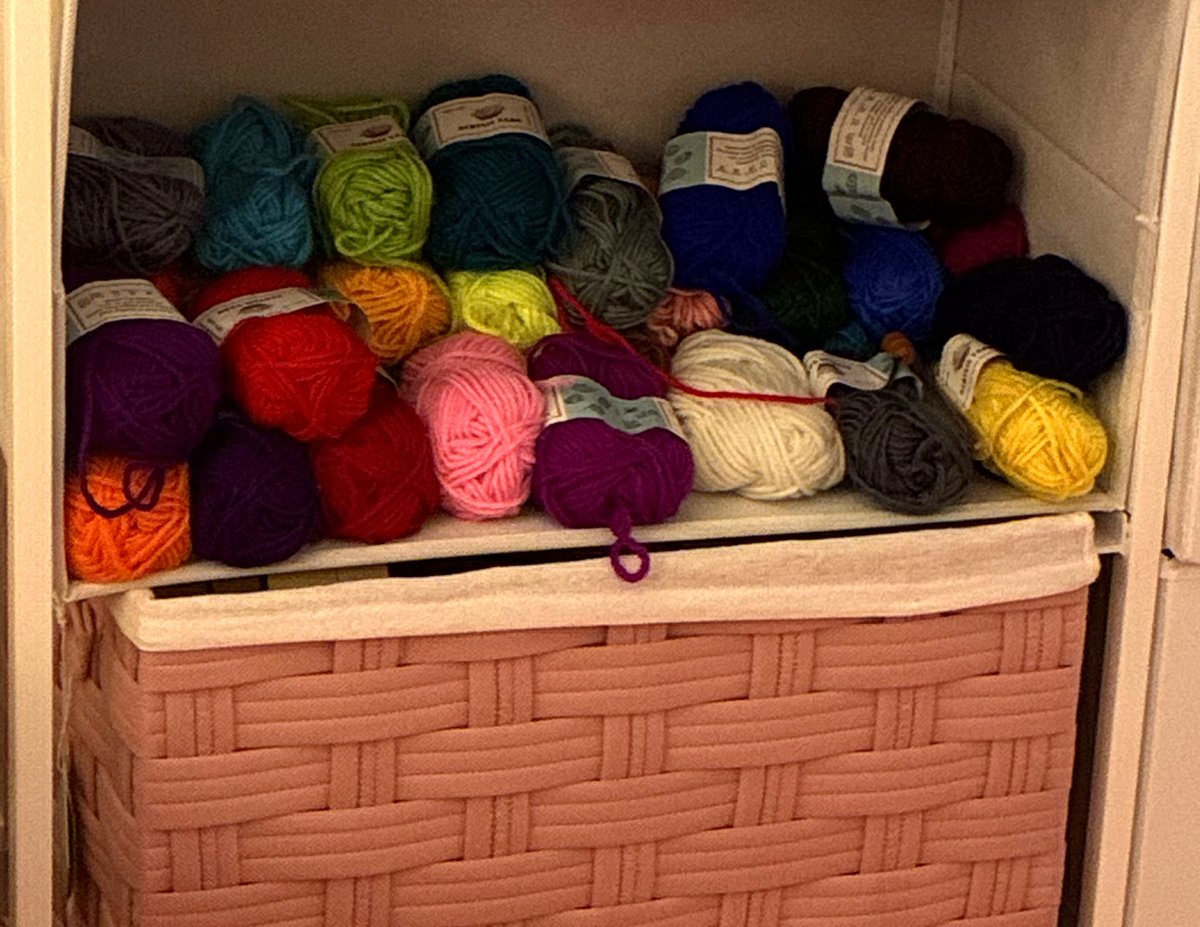 A shelf packed with colorful yarn skeins — purples, reds, pinks, blues, greens, and yellows — above a pink woven basket